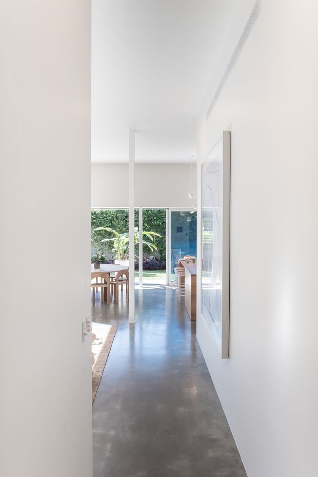 A bright, minimalist interior view looking down a hallway toward an open-plan dining and kitchen area. The space features smooth polished concrete flooring, white walls and ceiling, full-height glazing opening to a leafy garden, slim white structural posts, and simple timber dining furniture and cabinetry. A large framed artwork is mounted along the right wall.
