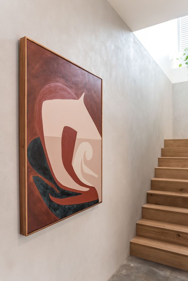 A contemporary interior stairway with warm timber steps rising beside a textured light plaster wall. A large framed abstract artwork with terracotta, cream, burgundy, and dark charcoal tones forms a feature on the wall. Natural light enters from a high opening with white louvres and a small plant visible near the upper right.
