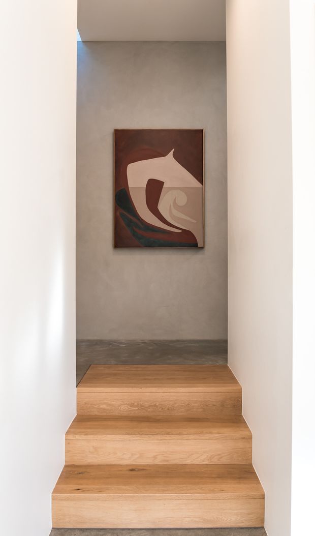 A minimalist stairway rises through a narrow white-painted opening to a small landing. The stairs and risers are finished in warm natural timber, while the landing has a dark grey polished or honed concrete-like floor and a softly textured grey wall displaying a framed abstract artwork. The composition is clean, restrained, and gallery-like, with strong vertical framing and warm wood contrasting against pale walls and grey surfaces.