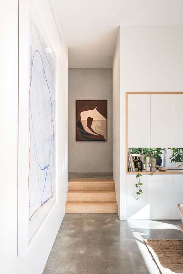 A bright minimalist interior hallway leading to a short set of oak stairs and a small landing. The space features polished grey concrete flooring, white walls and ceiling, a large abstract artwork, and built-in white cabinetry with warm timber framing and an open display niche containing books and plants. Natural light creates a clean, calm atmosphere.