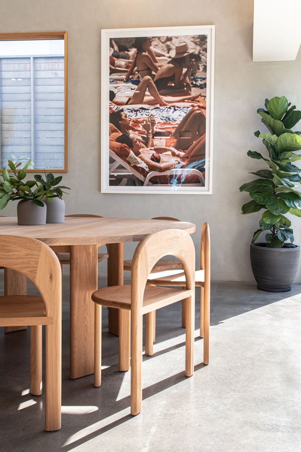 A sunlit dining area with a solid timber dining table and matching rounded-back timber chairs. The room has a smooth warm grey plaster wall, a polished concrete floor, potted greenery, a framed photographic artwork, and a timber-framed window with a translucent blind. Strong natural light creates long shadows across the floor.