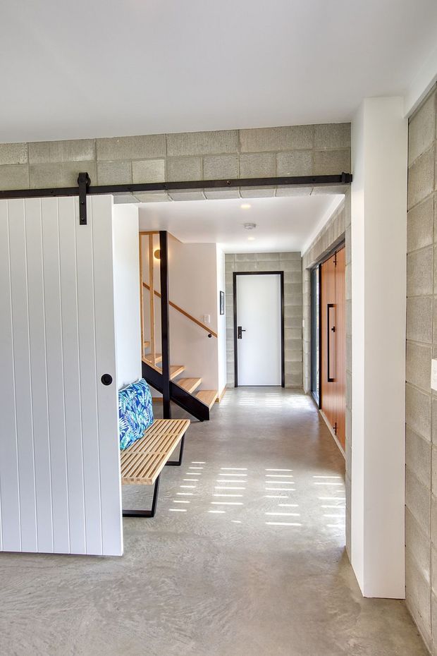 A bright contemporary entry hall with exposed concrete block walls, a polished concrete floor, and a white vertical-panel sliding barn door on a dark metal track. The corridor includes a timber bench with a colourful cushion, an open timber-and-black-metal staircase, and contrasting timber internal doors. Natural light creates striped shadows across the floor.