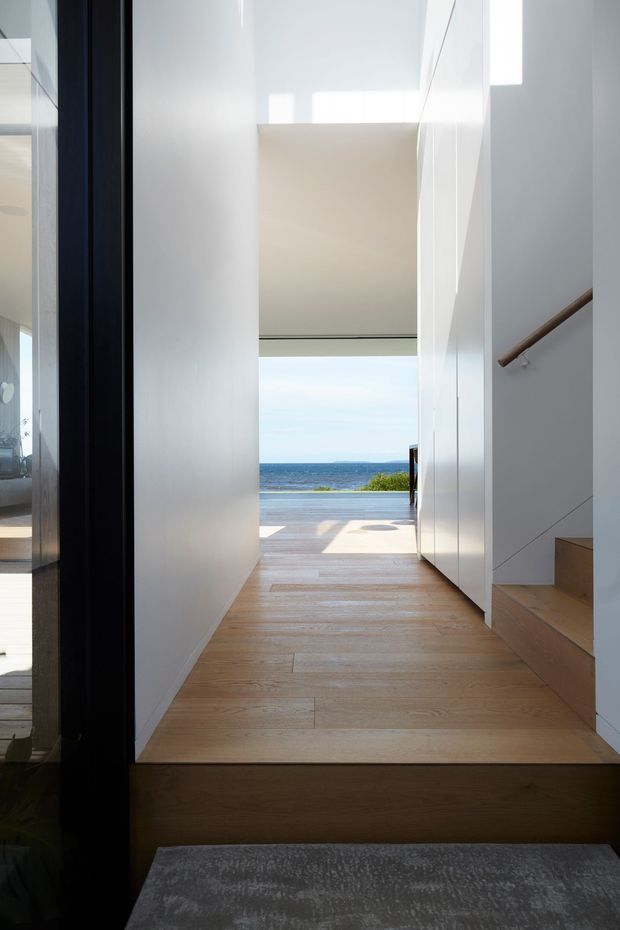 A bright minimalist interior hallway leading directly toward an ocean view and outdoor deck. The space features pale timber flooring, crisp white walls and built-in cabinetry, a black-framed glazed opening on the left, and a compact timber stair with a simple wood handrail on the right. Strong natural light creates a clean, airy transition between the interior and coastal landscape.