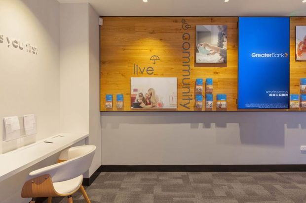A compact contemporary commercial reception or waiting area, likely in a bank or customer service office. The space features a white built-in counter with two molded chairs, a grey carpeted floor, and a warm timber feature wall with integrated shelving, promotional displays, and a large blue branded digital screen. White walls, recessed ceiling lights, and clean lines create a bright, professional environment.