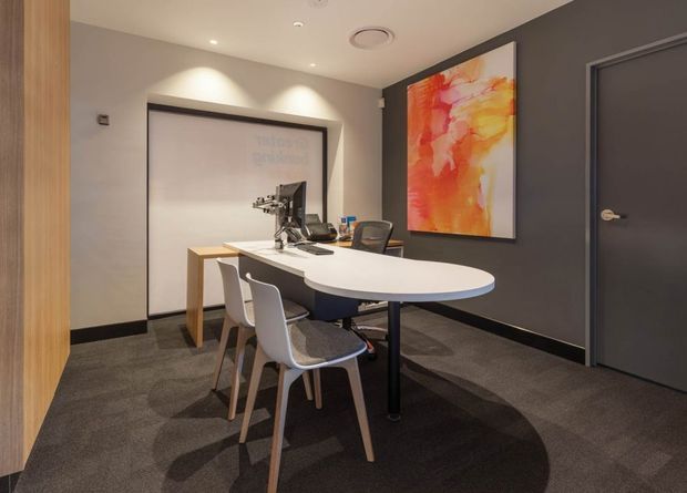 A compact contemporary office meeting room with a white oval conference table, mixed white and dark upholstered chairs, and computer equipment. Dark patterned carpet, charcoal grey walls and door, warm timber wall panelling, recessed downlights, and a vivid orange-red abstract artwork create a refined professional interior.