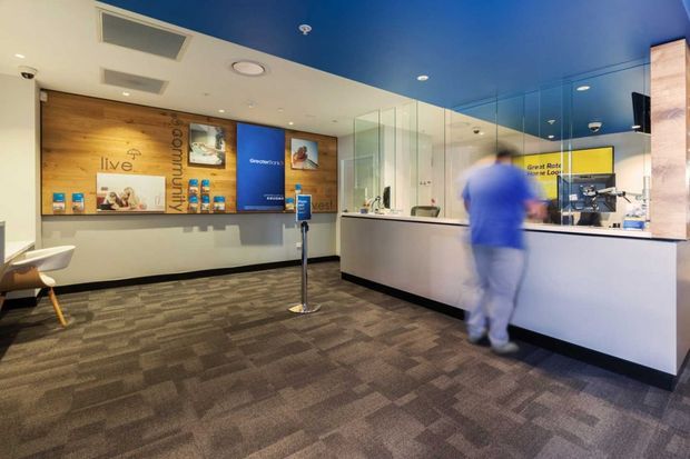 A modern commercial reception and service counter interior, likely a customer service or community-facing facility. The space features a long white counter with glass privacy screens, a blue ceiling, recessed lighting, patterned carpet flooring, timber wall panelling with integrated signage and display shelving, and branded informational graphics. A blurred person stands at the counter.