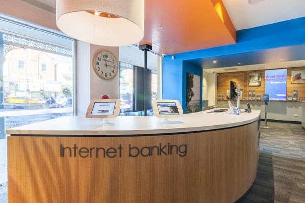 Bright modern commercial banking interior featuring a large curved internet-banking counter with vertical wood-grain cladding and a white solid-surface countertop. Tablet stations sit on the counter, with blue accent walls, warm timber ceiling detail, recessed lighting, large street-facing windows, and a carpeted floor. A clock and branded banking signage are visible in the background.