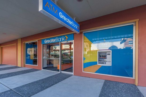 Exterior view of a Greater Bank branch with a blue ATM projecting from the facade, glazed entrance doors, a large blue window graphic, warm terracotta wall panels, and a patterned concrete forecourt beneath a covered canopy.