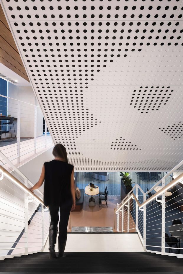 Interior view looking down a modern stairway in a commercial or public building. The staircase has dark treads, white cable railings, and white walls, while a dramatic perforated acoustic ceiling forms a patterned canopy overhead. A seating or lounge area is visible at the lower level, with contemporary lighting and blue-toned wall panels.