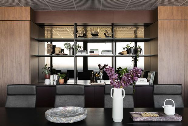 A refined contemporary office or study with a long dark desk, several black leather chairs, and a full-width built-in shelving and storage wall. Warm timber cabinetry frames open black metal shelving, which displays books, ceramics, plants, and decorative objects. The ceiling has an exposed linear metal grid, while large glazed panels behind the shelving bring in daylight. The palette is predominantly dark charcoal, black, warm wood, and muted neutral tones, with purple flowers adding a small accent.