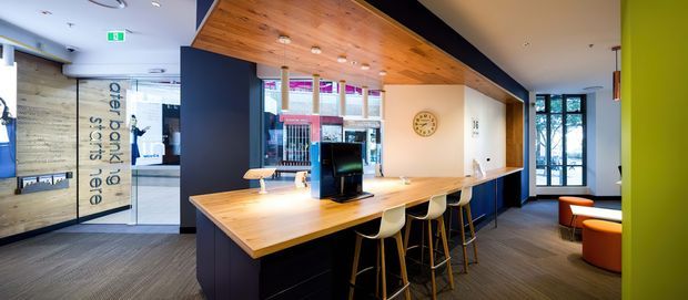 A bright contemporary commercial reception and shared work area. A long timber counter with integrated cabinetry and several bar stools sits beneath a warm timber-lined ceiling. The space includes a computer workstation, clock, dark blue joinery, carpeted flooring, glazed entry doors, and branded graphics on the adjacent glass partition.