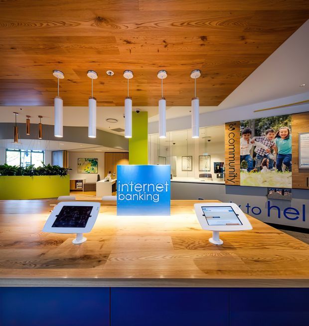 A bright, contemporary commercial banking interior with a large timber-clad ceiling and matching timber service counter. White cylindrical pendant lights hang above the counter, which displays tablet-based internet banking stations. The space includes lime-green planter and column accents, blue lower cabinetry, glazed partitions, framed artwork, and prominent customer-service graphics. The image is clearly a bank branch or financial services workspace.