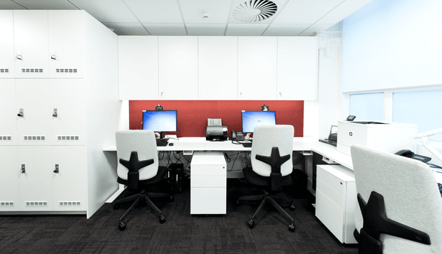 Bright, compact modern office workspace with white built-in storage lockers, a shared workstation for two, additional individual desk space, white ergonomic office chairs, computer monitors, printers, and a red feature wall. The room has a suspended white ceiling with recessed ventilation and dark carpet flooring, creating a clean, functional commercial interior.