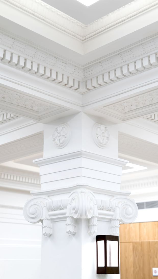 Close-up interior architectural detail dominated by an ornate white plaster or painted masonry column. The column features elaborate classical capitals with scrolls, floral rosettes, layered mouldings, dentil-like detailing, and decorative ceiling panels. A skylight or bright ceiling opening is visible above, with a dark-framed glazed doorway or partition and warm timber panelling in the background. The space appears highly decorative, bright, and heritage-inspired.
