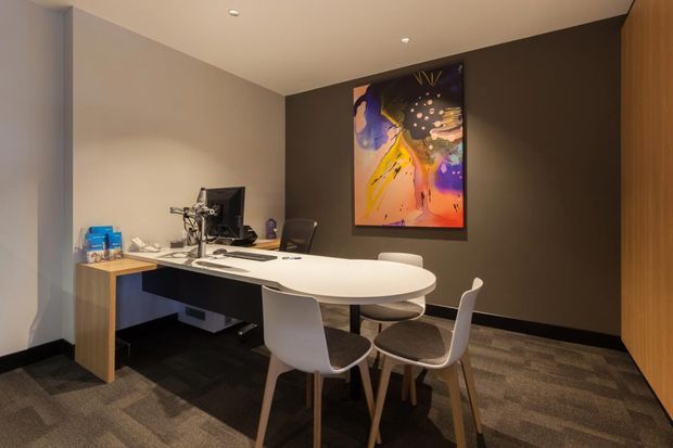 A compact, professionally fitted office or consultation room with a curved white meeting table, several contemporary chairs, a built-in work desk with computer equipment, dark feature wall, timber cabinetry, carpet tile flooring, recessed ceiling lights, and colourful abstract artwork.