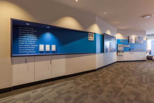A modern commercial interior corridor or reception area with a large blue graphic feature wall, integrated display panels, wall-mounted signage, and low built-in cabinetry. The space has recessed ceiling lights, neutral painted walls, dark skirting, and patterned grey-brown carpet. Promotional or informational graphics and a reception/display counter are visible toward the rear.