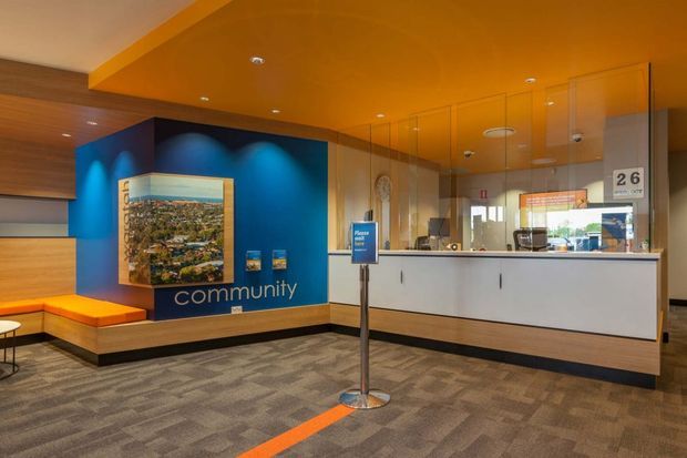 A contemporary commercial reception or service counter interior with a white built-in desk behind a glass security partition. The space features a vivid blue feature wall with the word “community,” an inset landscape image, orange ceiling and bench accents, recessed lighting, patterned carpet, and a queue marker with an orange floor line.