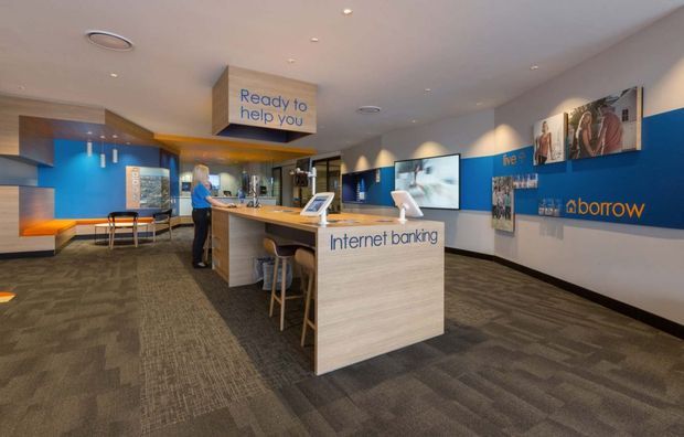 A contemporary bank branch interior with an open customer service and internet-banking area. The space features a central light-wood service island with stools and a freestanding transaction counter, blue branded feature walls, integrated digital screens, recessed ceiling lighting, and dark patterned commercial carpet. A small waiting or consultation area is visible at the rear.