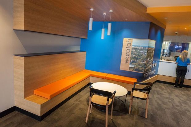 A brightly coloured contemporary commercial reception or waiting area with a built-in timber bench and orange upholstered seating wrapping around a small round table. The space features a vivid blue feature wall with signage and promotional graphics, pendant lights, carpeted flooring, and a reception counter visible in the background.