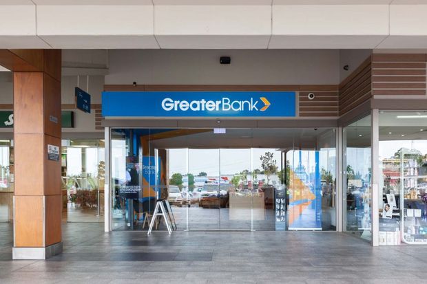 Front-facing view of a Greater Bank branch entrance within a contemporary commercial arcade. The glazed storefront has sliding glass doors, a bright blue bank sign, timber-clad columns and soffit details, horizontal timber slats, and large-format grey tiled flooring. Reflections and views through the glazing show the surrounding retail and outdoor context.