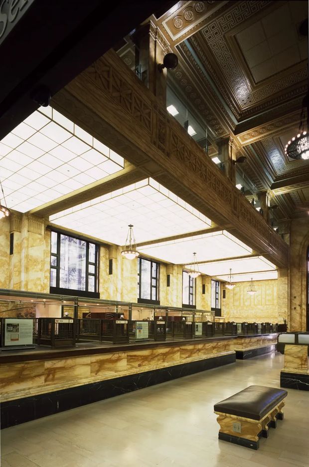 Grand historic commercial interior resembling a railway station or civic transit hall, with ornate neoclassical and Art Deco detailing. The space features richly patterned coffered ceilings, decorative friezes, marble-clad walls and counters, dark trim, large illuminated ceiling panels, chandeliers, tall windows, and a long bank of ticket or service counters. A substantial decorative balcony or mezzanine runs across the upper level.