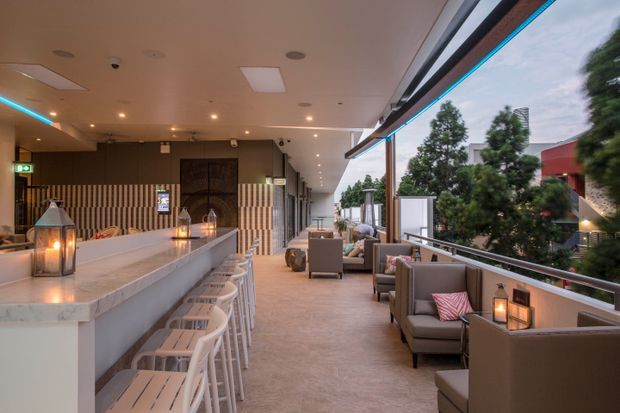 A covered open-air hospitality terrace with a long marble-look bar and white stools on the left, and upholstered lounge seating along the glazed edge on the right. Warm lanterns, recessed ceiling lights, and blue concealed accent lighting create an inviting evening atmosphere. The terrace overlooks landscaped suburban surroundings and includes retractable or sliding overhead glazing.