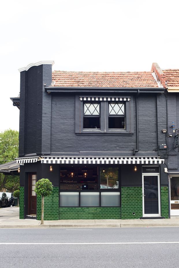 Street-facing facade of a compact two-storey commercial building, likely a hospitality venue. The exterior combines dark painted brickwork, a terracotta tiled pitched roof, green glazed brickwork at ground level, black-and-white striped awnings, diamond-patterned upper windows, and a glazed entrance door. The image is photographed from across the street.