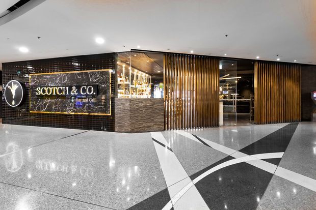 A polished commercial restaurant entrance with a prominent illuminated marble-look sign reading “SCOTCH & CO. Bar and Grill.” The sophisticated interior features dark tiled walls, warm timber vertical slats, a textured stone-clad host or bar counter, glass openings into the venue, recessed ceiling lighting, and a geometric black, white, and grey polished floor pattern.