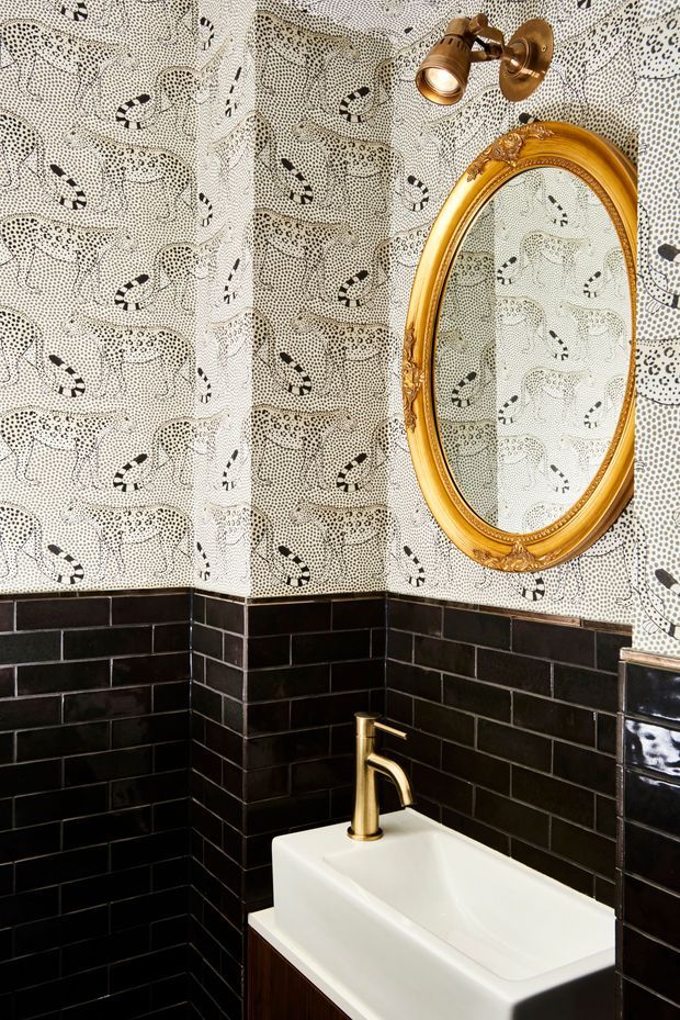 A compact, dramatically styled bathroom corner featuring glossy black subway wall tiles, a white vessel basin with a brass-gold tap, patterned monochrome wallpaper, an ornate oval gold-framed mirror, and a brass wall spotlight. The eclectic decorative treatment creates a bold, vintage-inspired interior.