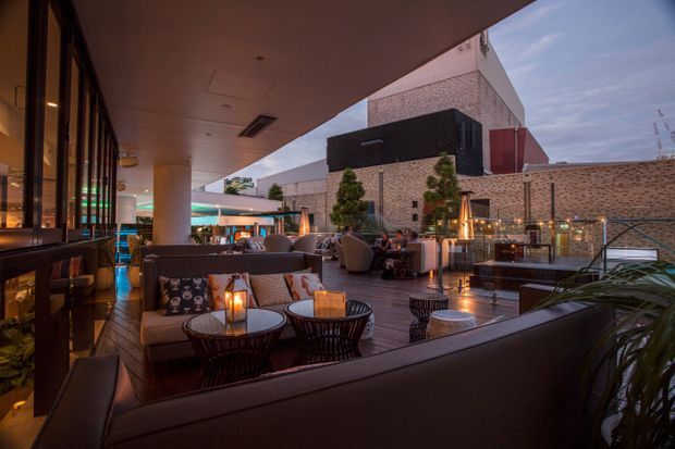 A sophisticated rooftop or elevated outdoor lounge at dusk, furnished with sofas, armchairs, low round tables, lanterns, and planters. The space is adjacent to a glazed commercial interior on the left and enclosed by contemporary brick and metal architectural elements, with warm ambient lighting and city-facing openness.