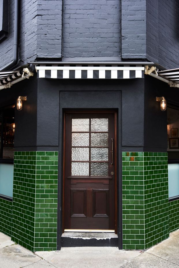 Street-level view of a narrow commercial entrance framed by dark painted masonry and green glazed wall tiles. A dark timber door with textured glass sits beneath a black-and-white striped awning, with warm wall-mounted lanterns and partial interior bar or shop areas visible at either side.