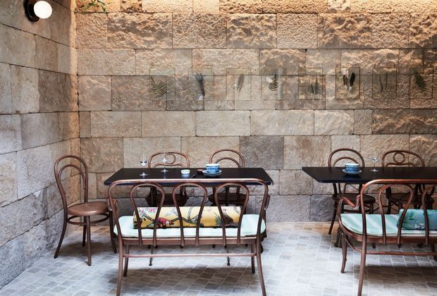 A rustic café or dining space with exposed rough-cut stone walls, a pale tiled floor, and two dark rectangular dining tables. Bentwood-style metal chairs and bench seating surround the tables, with small cups and table settings. Decorative botanical artwork is mounted across the stone wall, and a wall light adds warm illumination.