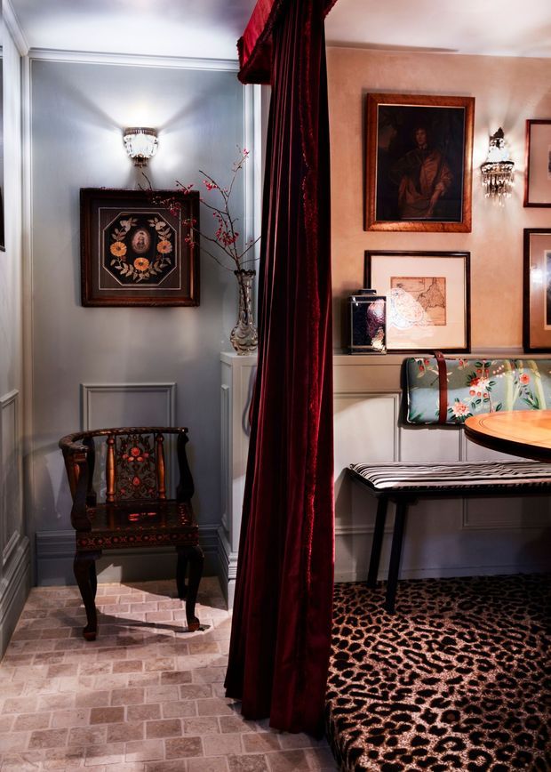 Eclectic, richly decorated interior vignette divided by a floor-length burgundy velvet curtain. The left alcove has pale blue-grey paneled walls, a decorative wall sconce, framed artwork, an ornate dark wood chair, a tall vase, and a light brick-pattern tiled floor. The right side features warm beige walls with framed artwork, crystal wall sconces, a built-in upholstered bench with floral cushions, a round timber table, a striped seat, and a dark animal-print carpet. The overall atmosphere is theatrical, vintage, and highly ornamented.