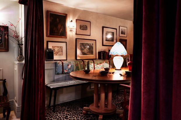 A richly decorated traditional dining area viewed through deep burgundy curtains. The room features a round polished wood dining table, upholstered dining chairs, a patterned bench, framed artwork arranged across warm neutral walls, a glowing decorative table lamp, and a dark patterned floor covering. White panelled wainscoting and layered textiles contribute to an intimate, theatrical atmosphere.