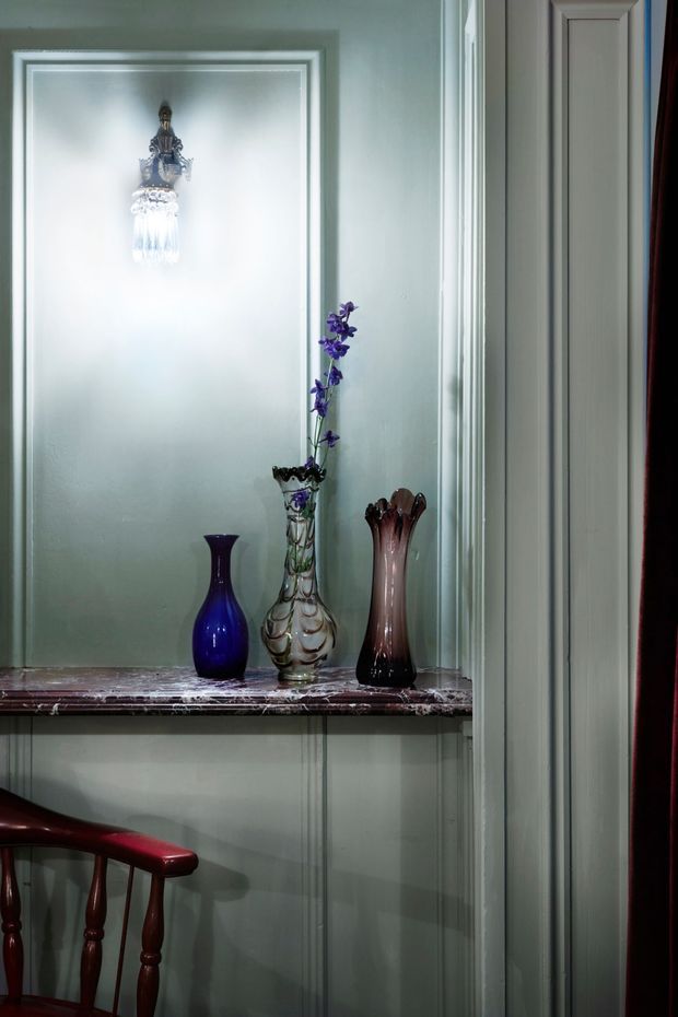 A narrow, traditionally detailed interior alcove or hallway niche with pale sage-green painted wall panelling and ornate white moulded trim. A dark polished marble shelf displays three decorative glass vases, with a small crystal wall sconce above. Part of a dark red wooden chair and a deep burgundy curtain are visible at the edges, contributing to a refined, heritage-inspired atmosphere.