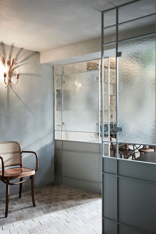 A refined interior passage or entry area with pale plaster walls, a grey metal-framed glazed partition featuring textured translucent glass, and a matching hinged door. The space has a light brick-paver floor, a woven cane and timber armchair, and a sculptural wall sconce. A dining area is partially visible through the partition. The overall look is warm, understated, and vintage-influenced with strong industrial steel-and-glass detailing.