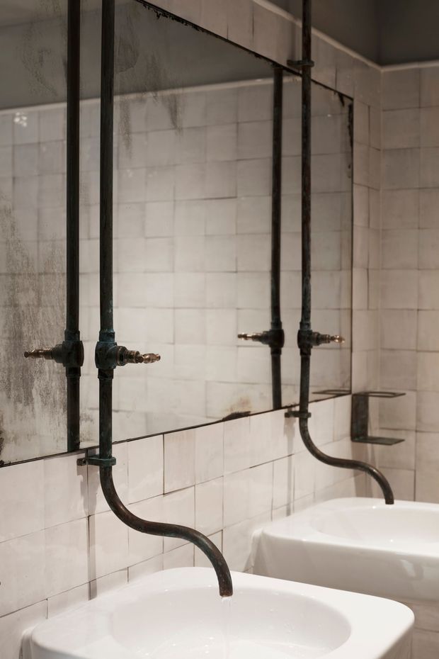 Close-up view of a bathroom with two white wall-mounted basins, exposed dark metal plumbing and taps, and a large frameless mirror reflecting the fixtures. White rectangular wall tiles and aged industrial pipework create a raw, utilitarian atmosphere.