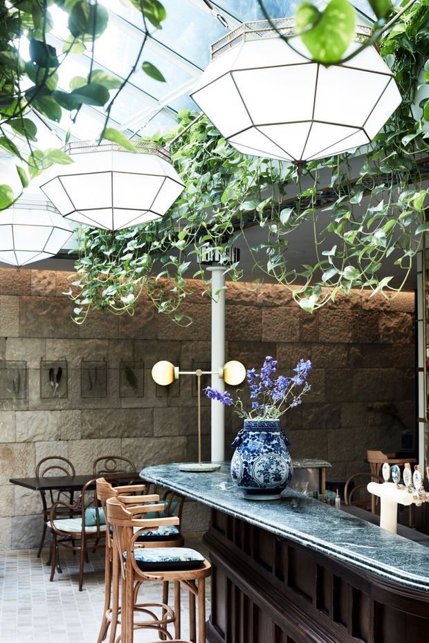 A refined indoor dining and bar area with a long dark wood bar, polished grey stone benchtop, and timber bar stools. A textured natural stone wall forms the backdrop, while lush trailing greenery hangs beneath a glazed roof or atrium. Large geometric white pendant lights illuminate the space, complemented by a blue-and-white ceramic vase and patterned upholstery.