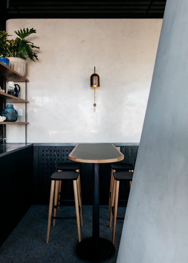 A compact, dark-toned café-style dining nook with a small rounded rectangular bar table and stools, set against a textured pale plaster wall. Black timber wall panelling, open shelving with plants and ceramics, a suspended pendant light, and a dark speckled floor create a refined, contemporary interior.