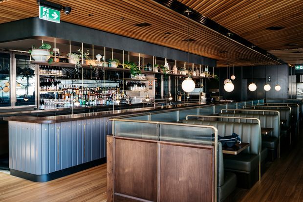 A stylish contemporary bar and dining venue with a long backlit bar, extensive bottle display, upholstered booth seating, and pendant globe lights. The space features dark timber and black finishes contrasted with warm timber slatted ceilings and flooring, with metal and glass booth partitions.