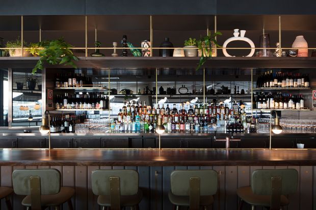 A sophisticated bar interior with a long dark timber counter, upholstered bar stools, illuminated liquor shelving, mirrored backsplashes, brass accents, glassware, and decorative shelving displaying plants and ceramics. The space has a warm, moody hospitality atmosphere with refined contemporary detailing.