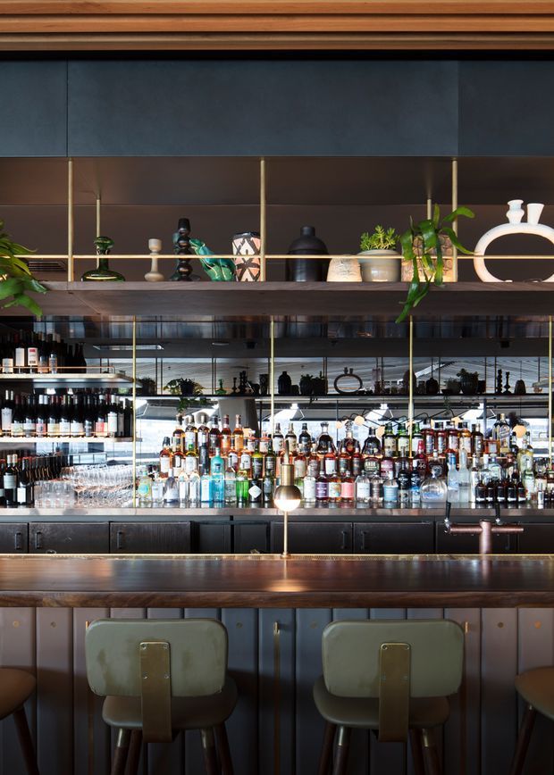A richly detailed bar interior with a long dark timber counter and upholstered bar stools in the foreground. Behind the counter are extensive backlit shelves filled with bottles, glassware, and decorative objects, framed by dark cabinetry, mirrored panels, brass-toned vertical supports, and timber shelving. The space has a moody, sophisticated hospitality fit-out with layered lighting and abundant material detail.