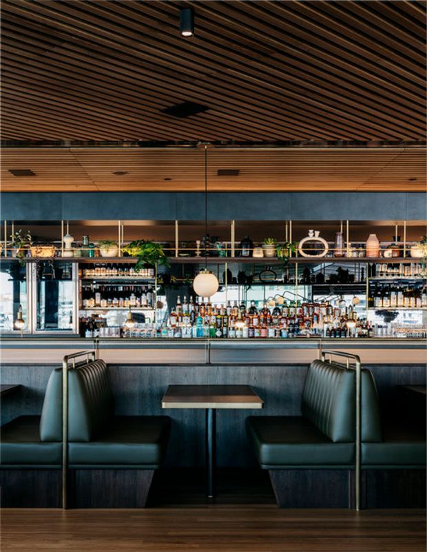 A sophisticated bar and dining area with upholstered booth seating, a small central table, and a fully stocked back bar reflected in a large mirror. The space features dark timber slatted ceilings, dark wood finishes, blue-grey wall panelling, metallic detailing, glass shelving, and warm globe lighting. The overall atmosphere is moody, refined, and contemporary, resembling a restaurant or cocktail bar interior.