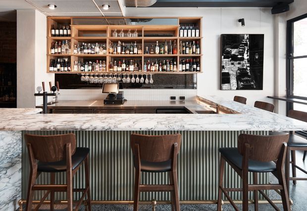 A sophisticated commercial-style bar and dining space with a large marble-topped island, fluted vertical-panelled bar front, dark timber cabinetry, and extensive open shelving displaying bottles and glassware. Three timber bar stools sit at the island, while a back counter, espresso machine, tiled splashback, framed artwork, and large windows complete the refined interior.