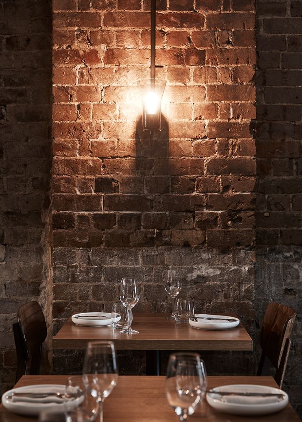 A moody restaurant dining interior with exposed, weathered red brick walls, warm wall-mounted lighting, and timber dining tables set with white plates, cutlery, and wine glasses. Dark wooden chairs and intimate table spacing create an industrial, rustic atmosphere.