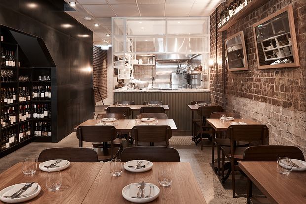 A compact industrial-style restaurant dining room with closely arranged timber tables and dark wood chairs, set with plates, cutlery and glassware. Exposed brick walls, black metal cabinetry and shelving, polished concrete flooring, and a glazed open kitchen at the rear define the space. Warm pendant and track lighting create an intimate atmosphere.