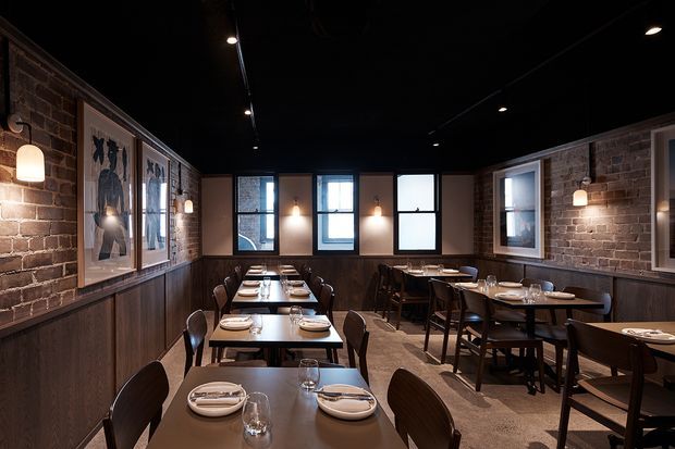 A dimly lit brick-walled dining room or restaurant interior with dark timber wainscoting, rows of neatly set wooden tables and chairs, framed artwork, exposed brick walls, pendant sconces, and black ceilings with recessed track lighting. The space has a warm, refined industrial atmosphere.