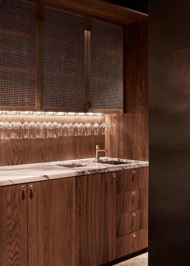 A compact, richly detailed home bar or drinks station with dark walnut cabinetry, a white marble countertop with grey veining, integrated sink and brass-toned faucet. Hanging wine glasses are displayed beneath textured mesh-front overhead doors, with warm under-cabinet lighting creating an intimate, luxurious atmosphere.
