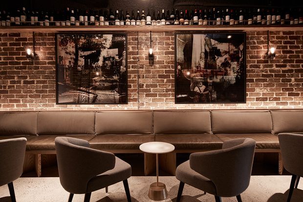 A warmly lit, intimate bar or lounge interior with an exposed red-brick feature wall, dark shelving displaying bottles, framed artwork, a long brown upholstered banquette, and sculptural lounge chairs around a small round table. The space has a refined, moody hospitality atmosphere with strong industrial architectural detailing.