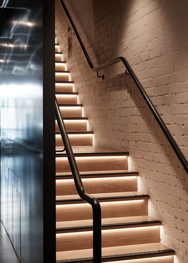 A close-up view of an interior stairway with warm timber treads, integrated step lighting, dark metal handrails, and exposed painted brick walls. A reflective dark metal-clad wall runs along the left side, creating a refined industrial atmosphere.